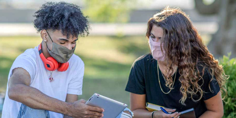 Two students talk on a college campus while wearing masks.