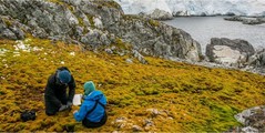 Two researchers in jackets sit on a yell-colored moss ground; water is in the background
