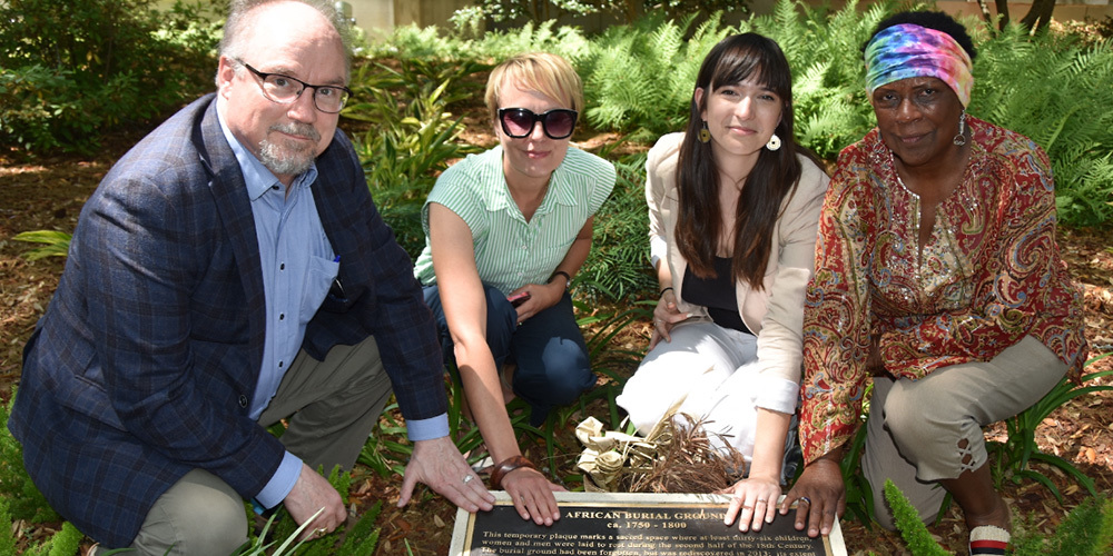 Four people touch a plaque on the ground commemorating a burial site.