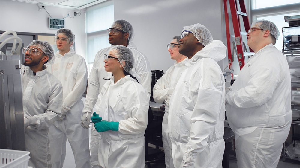 A group of students in white lab gear look on as a researcher makes a demonstration.