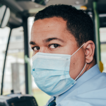 Photo shows a Hispanic/Latino bus driver wearing a facemask. 