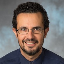 Portrait of Dr. Matias Valenzuela. He has short, curly hair, a sparse beard, and wears glasses. 