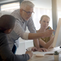 A man in a dress shirt pointing to a computer monitor while a man in a black shirt and a woman in a brown shirt are looking at the monitor.
