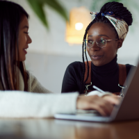 Woman in a black shirt sitting and looking at a girl in a white shirt, who is using a laptop.