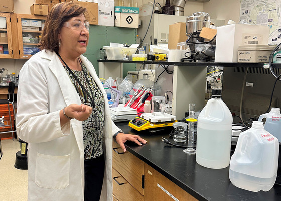 Woman in white lab coat gestures while standing in a university laboratory