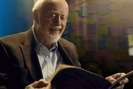 Elderly man with white beard poses in front of collage of Post-it Notes