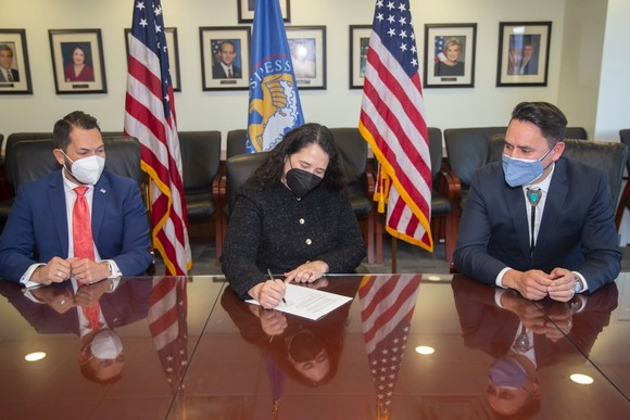Three people sitting at a table. The SBA Administrator is signing a document. The American and SBA flags are in the background.