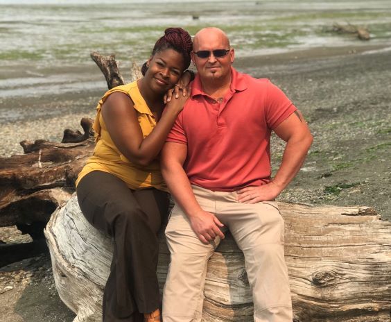 Makieda and Reno Hart sit on a log, a beach and body of water is in the background