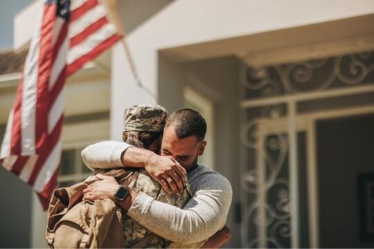 A woman in military uniform is warmly embraced by a man at the entrance of a home, with a U.S. flag hanging in the background.