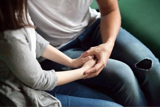 Two people sitting side by side, holding hands. A woman’s hands rest on a man’s leg as he gently holds them.