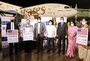 Seven people, three of whom are holding signs with the U.S. flag and COVAX, stand on the tarmac in front of crates of vaccine doses and a jet. 