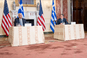 Secretary Blinken and Greek Foreign Minister Dendias sit at separate desks in a formal meeting room, with their countries’ flags behind them.