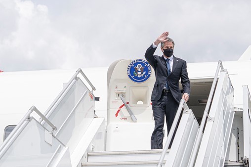 Secretary Blinken, wearing a grey suit, waves as he steps out of a jet and heads to the stairs down to the tarmac.