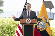 Secretary Blinken stands at a podium outdoors, with the flags of the USA and Ecuador behind him. 