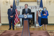 Secretary Blinken standing at a podium and Ambassadors Moore and Uyhara behind him in the Benjamin Franklin Room. 
