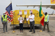 Four officials standing on an airport tarmac with a pallet of Vaccines with the United States and Senegal flag. 