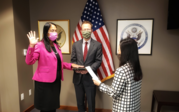 Ambassador Sison in a pink blazer and black dress being sworn in front of an American flag and two Department of State officials. 