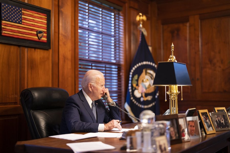 President Biden speaks on the phone as he sits at a desk in a wood-paneled office. 