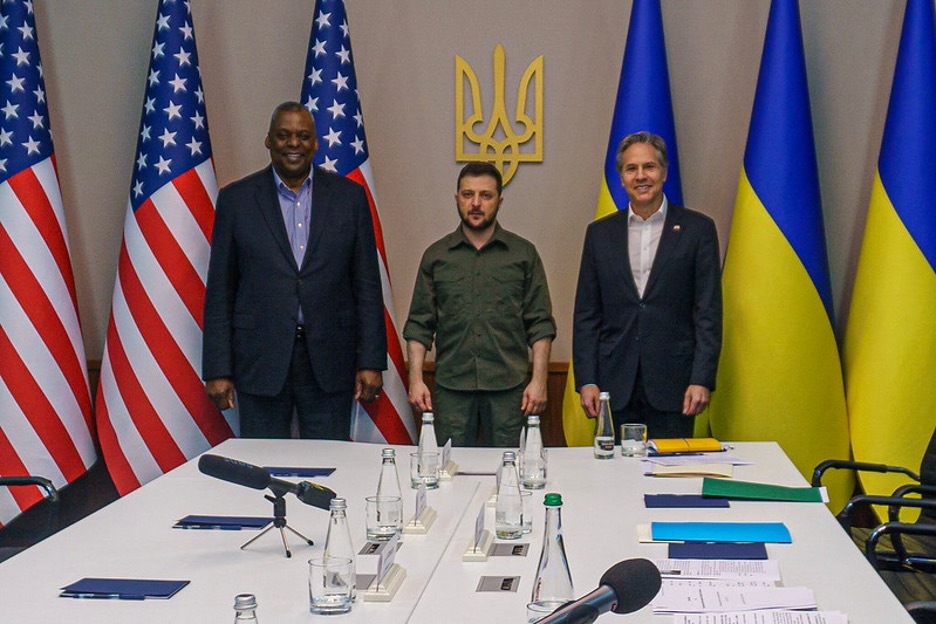 Secretary Austin and Secretary Blinken smile as they stand on either side of Ukrainian president Zelenskyy, with their country flags behind them. 