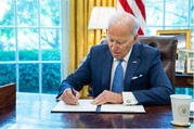 President Biden, wearing a blue suit and tie, signs a document at his desk in the oval office. 