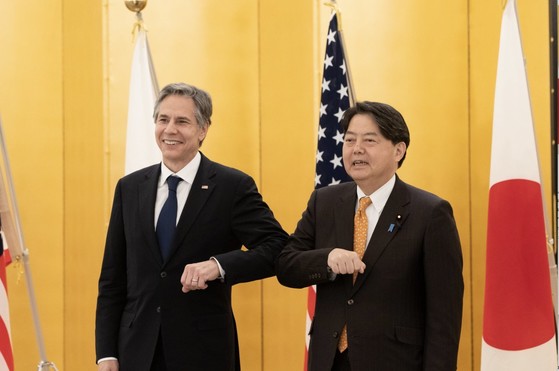 Secretary Blinken and Japanese Foreign Minister Hayashi stand in front of their country flags and smile as they bump elbows. 