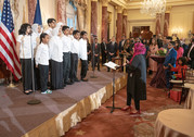 In an ornate reception room, a woman stands in front of a small stage and directs a small children's choir wearing white dress shirts. 