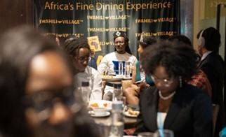  Members of the African Diaspora Youth and Exchange Alumni eating in front of a black and orange banner that reads: “Africa’s Fine Dining Experience”.