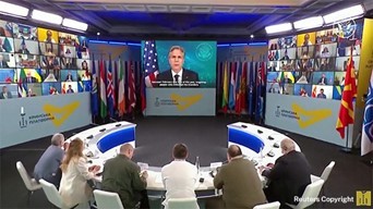 Secretary Blinken giving remarks via a large monitor, with other participants on screens and Six people are seated at table facing the screens.