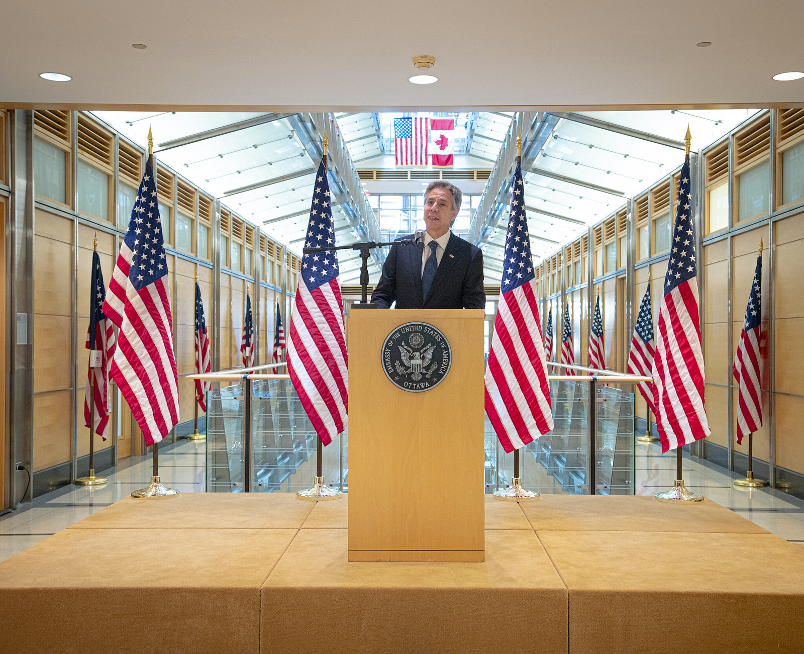 Secretary Blinken speaking in front of U.S. flags at the podium with the U.S. and Canadian flags hanging side by side in the foreground. 