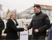 Ambassador Brink shakes hands with a man outside as they stand in front of a USAID delivery of generators. 