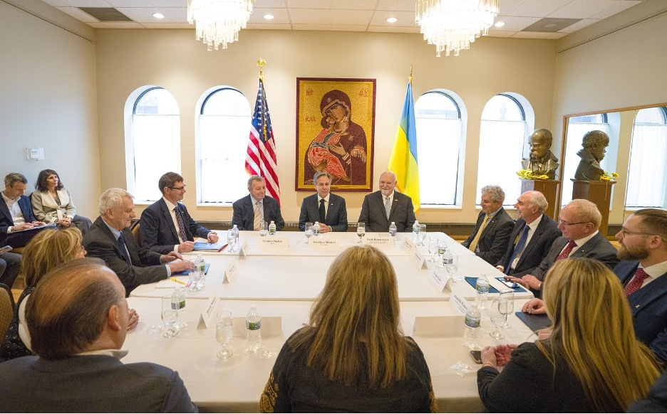 Secretary Blinken and 12 people sit around a square table. Behind the Secretary is a large religious painting, and the U.S. and Ukrainian flags.