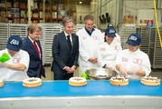 Secretary Blinken stands with workers in white aprons and blue hats who are decorating cheesecakes.