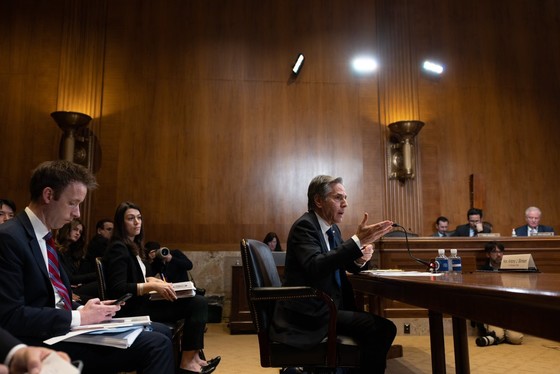 Secretary Blinken sits at a wooden table in a wood-paneled room, with others, also dressed in suits, sitting in the room. 