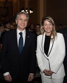 Secretary Blinken and Foreign Minister Joly, both wearing suits, stand next to each other and smile. Other people are in the background. 