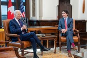 President Biden and Canada Prime Minister Trudeau are seated and talking in an official office. U.S. and Canada flags are in the background.