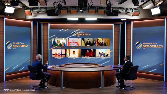 President Biden and Secretary Blinken sit at a large, round table and address other world leaders on screens projected in front of them. 