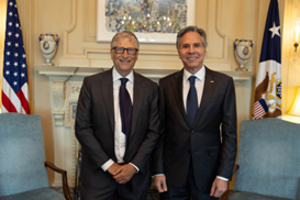  Secretary Blinken and Bill Gates, both wearing suits and smiling, stand in front of a fireplace in a formal room. 