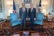 Secretary Blinken and Deputy Secretary Verma, both wearing suits and both smiling, stand in front of two light blue chairs in a formal room. 