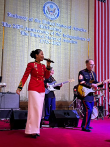 People perform music on stage. In the background, a sign notes the 247th anniversary of US independence. 