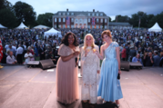 Three women in long dresses (in pale shades of red, white, and blue) stand on a stage in front of a large outdoor crowd. 