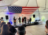 Five musicians perform on a stage under a white tent, with a large flag of the United States above them.