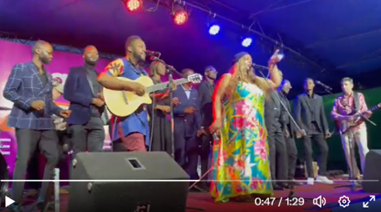 A large group of musicians, including a woman in a floral dress, perform on stage. 