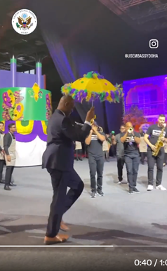 The ambassador marches with a colorful umbrella while a group of musicians play brass instruments. 