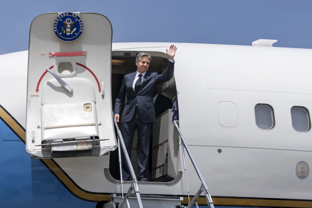 Secretary Blinken smiles and waves at the open door of a jetplane, with a sunny blue sky behind him.