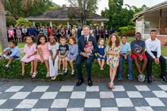 Secretary Blinken, holding a baby, sits with children on a low wall outside. A checkerboard-patterned patio is in the foreground. 