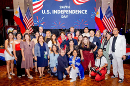 About 20 people, many wearing red, white, and blue, pose for a photograph on a stage while celebrating the Fourth of July. 