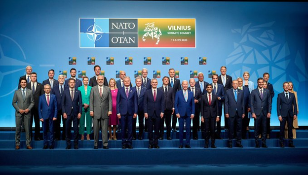 NATO members stand in three rows on blue carpeted steps in front of a blue background with the NATO Summit logo. 