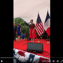 A woman in a red dress stands on a stage and sings. Behind her are the flags of France and the U.S.