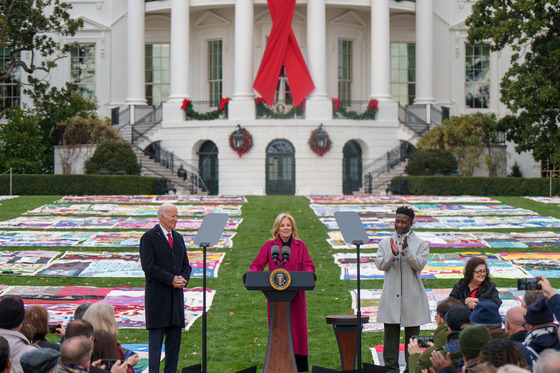 Alt text: First Lady Jill Biden speaks at a podium in front of the White House and the AIDS Memorial Quilt.