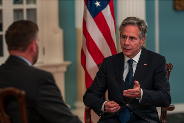 Secretary Blinken sits in a chair, speaking, with U.S. flag displayed behind him (State Department/Chuck Kennedy).
